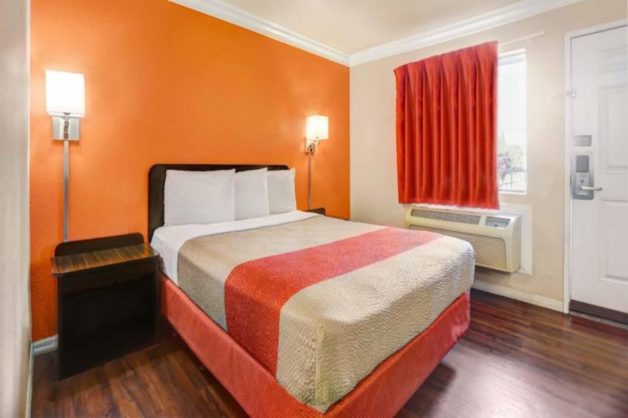 A hotel room with a bed, nightstand, wall-mounted lamps, an air conditioning unit, and a door.