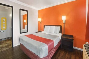 Una habitación de hotel con cama, mesita de noche, espejo y una pared con interruptor de luz.