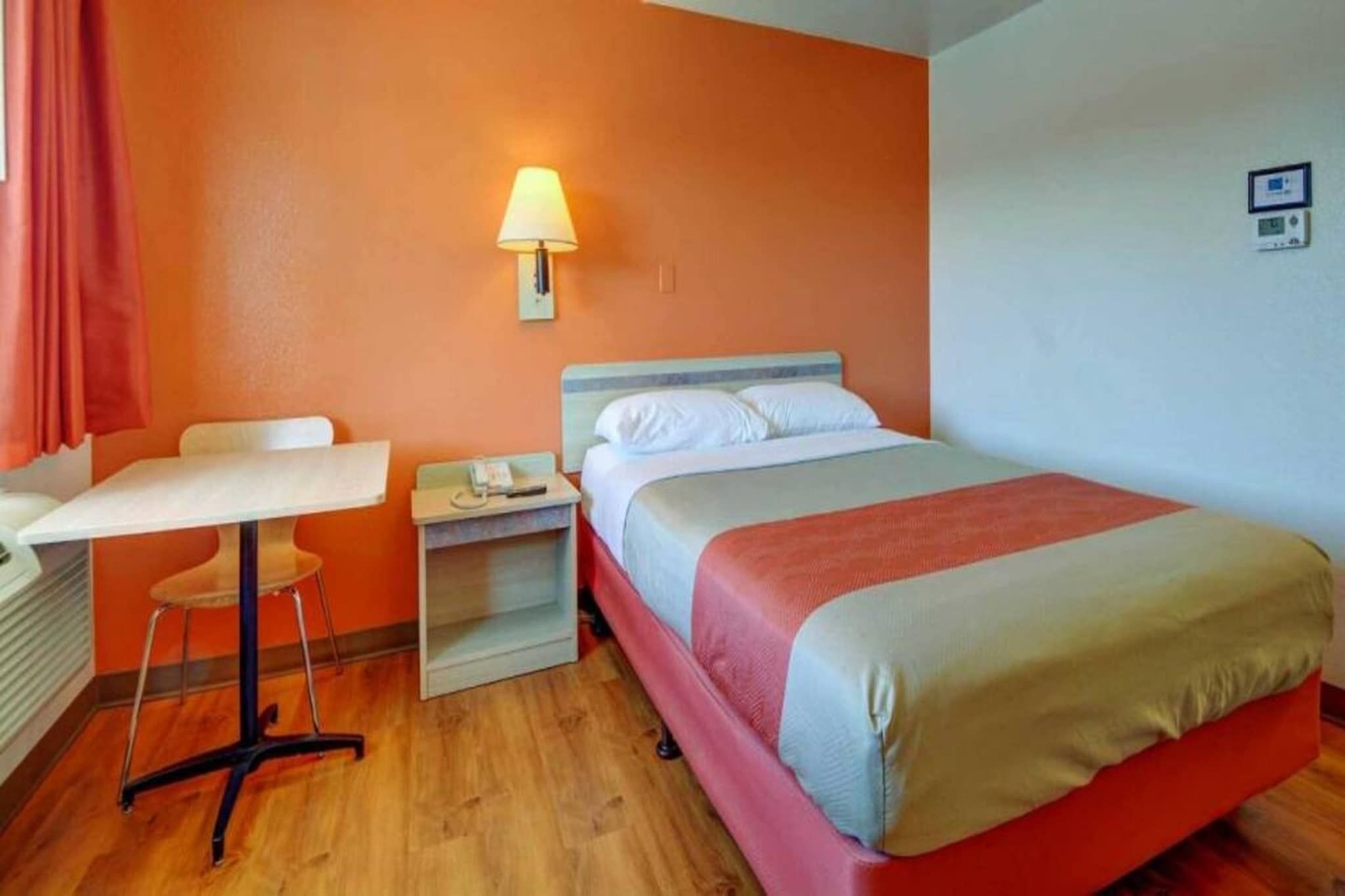 A hotel room with a bed, a desk, a chair, a nightstand, and a wall-mounted lamp.