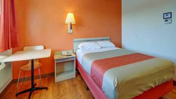 A hotel room with a bed, a desk, a chair, a nightstand, and a wall-mounted lamp.