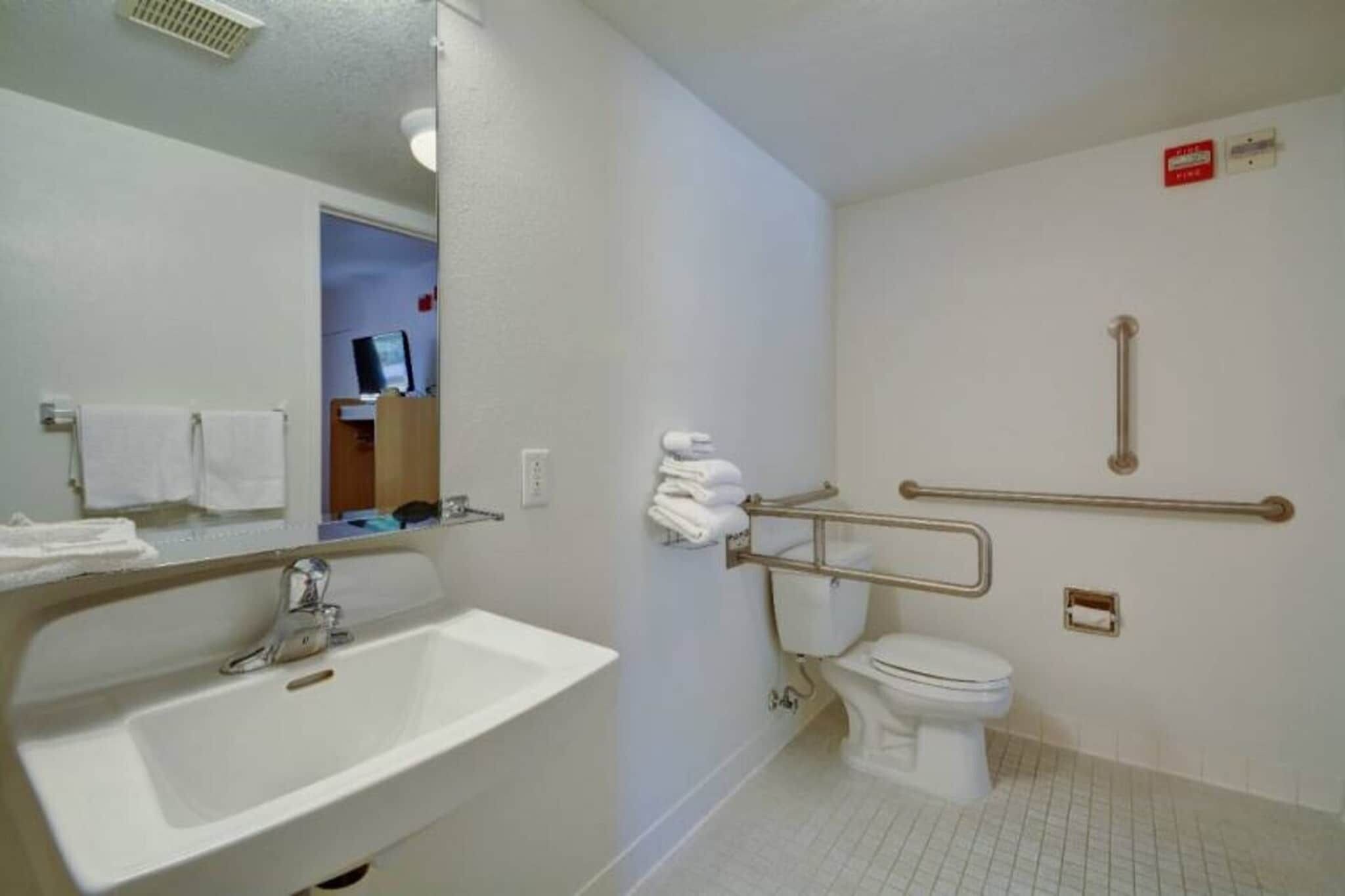 Une salle de bain d’hôtel standard, équipée d’un lavabo, de toilettes et de barres d’appui.