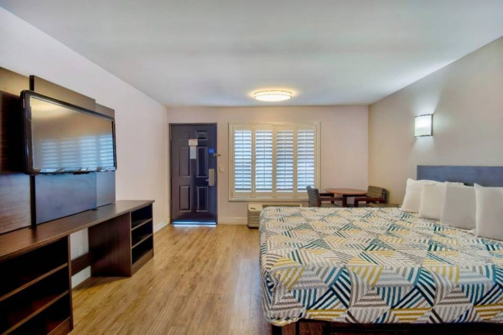 A bedroom with a bed, a desk, a chair, a TV, and a window with blinds.