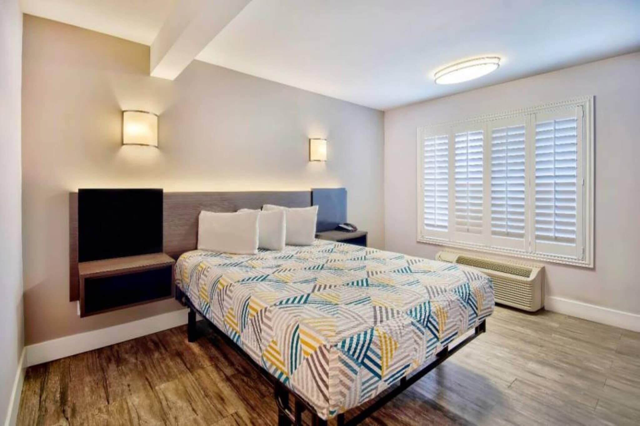 A bedroom with a bed, bedside tables, a wall-mounted TV, a window with blinds, and a built-in heater.