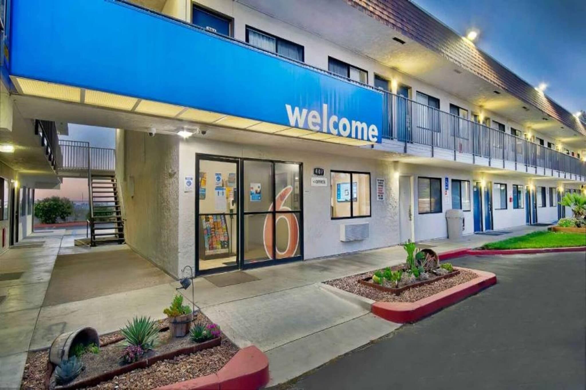 A hotel with a blue 'Welcome' sign, a covered entrance, and a landscaped area with plants.