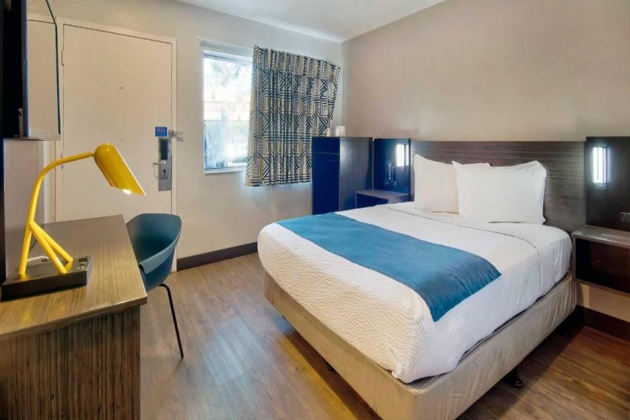 A hotel room with a bed, bedside tables, a desk with a lamp, a chair, and a window with patterned curtains.
