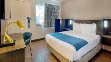 A hotel room with a bed, bedside tables, a desk with a lamp, a chair, and a window with patterned curtains.