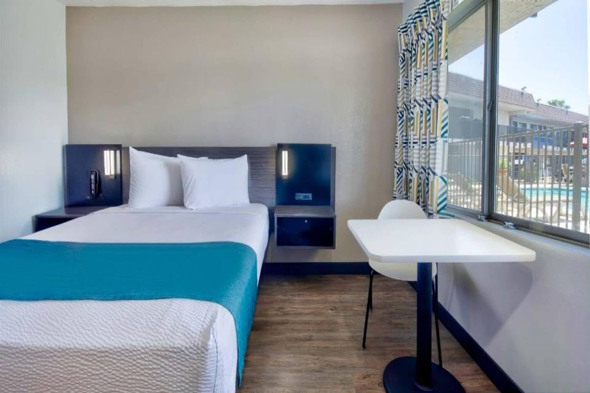 A hotel room with a bed, bedside tables, a desk, and a window with a view of a pool.