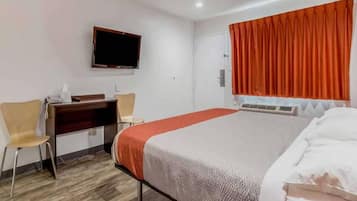 A hotel room with a bed, a desk, two chairs, a television, and an air conditioning unit.