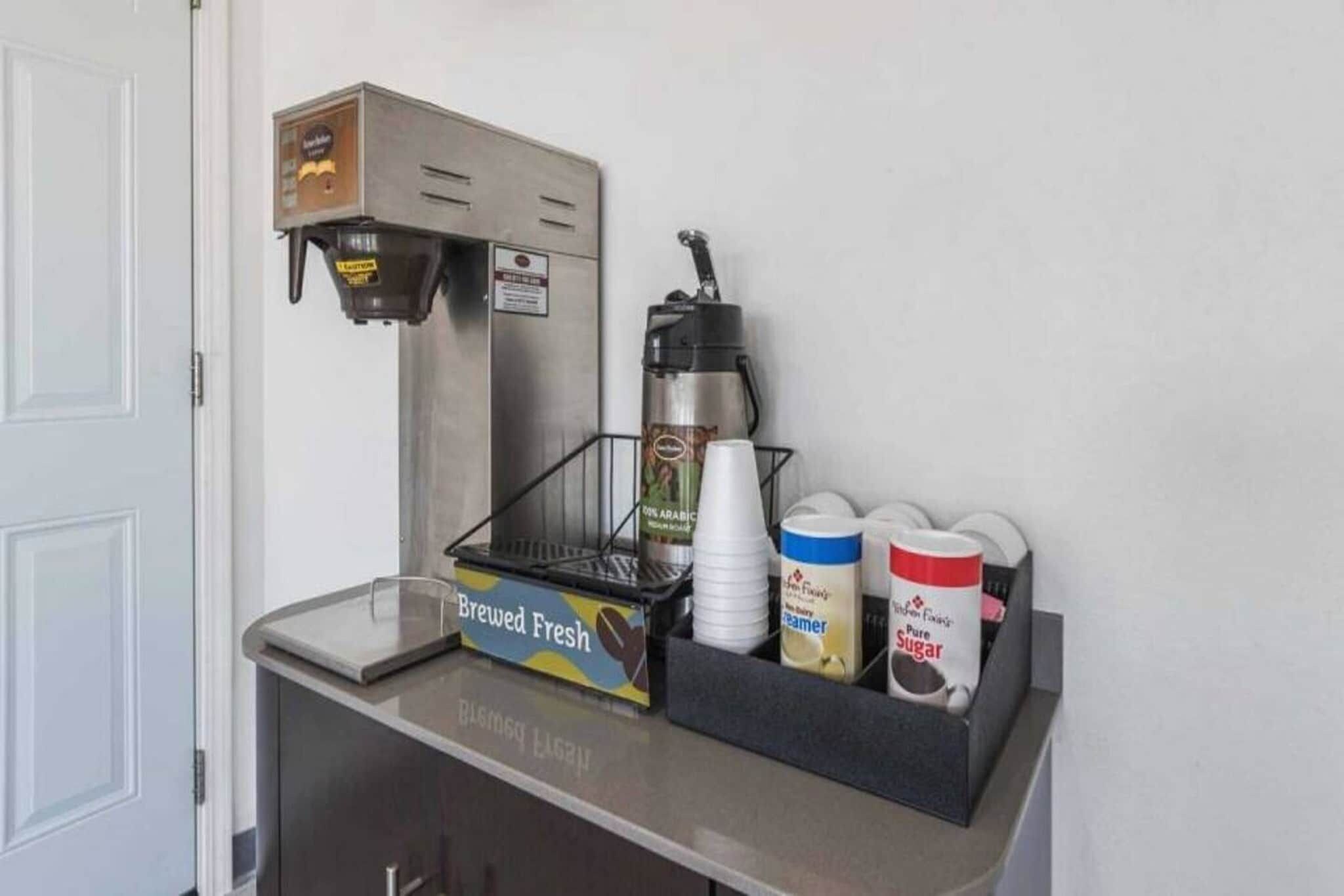 Una cafetera, un portavasos para tazas de café, y recipientes con leche condensada y azúcar sobre una encimera.