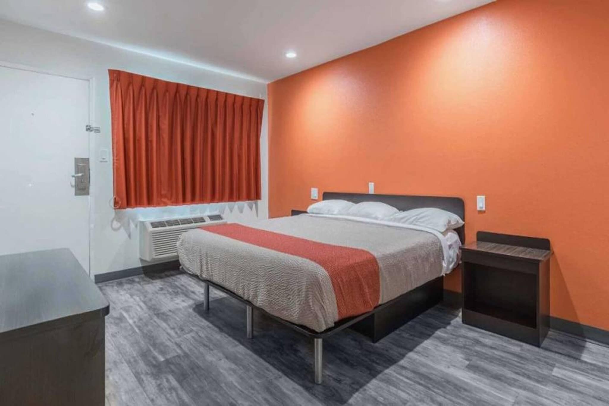 A hotel room with a bed, a nightstand, a desk, a window with curtains, and an air conditioning unit.