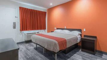A hotel room with a bed, a nightstand, a desk, a window with curtains, and an air conditioning unit.