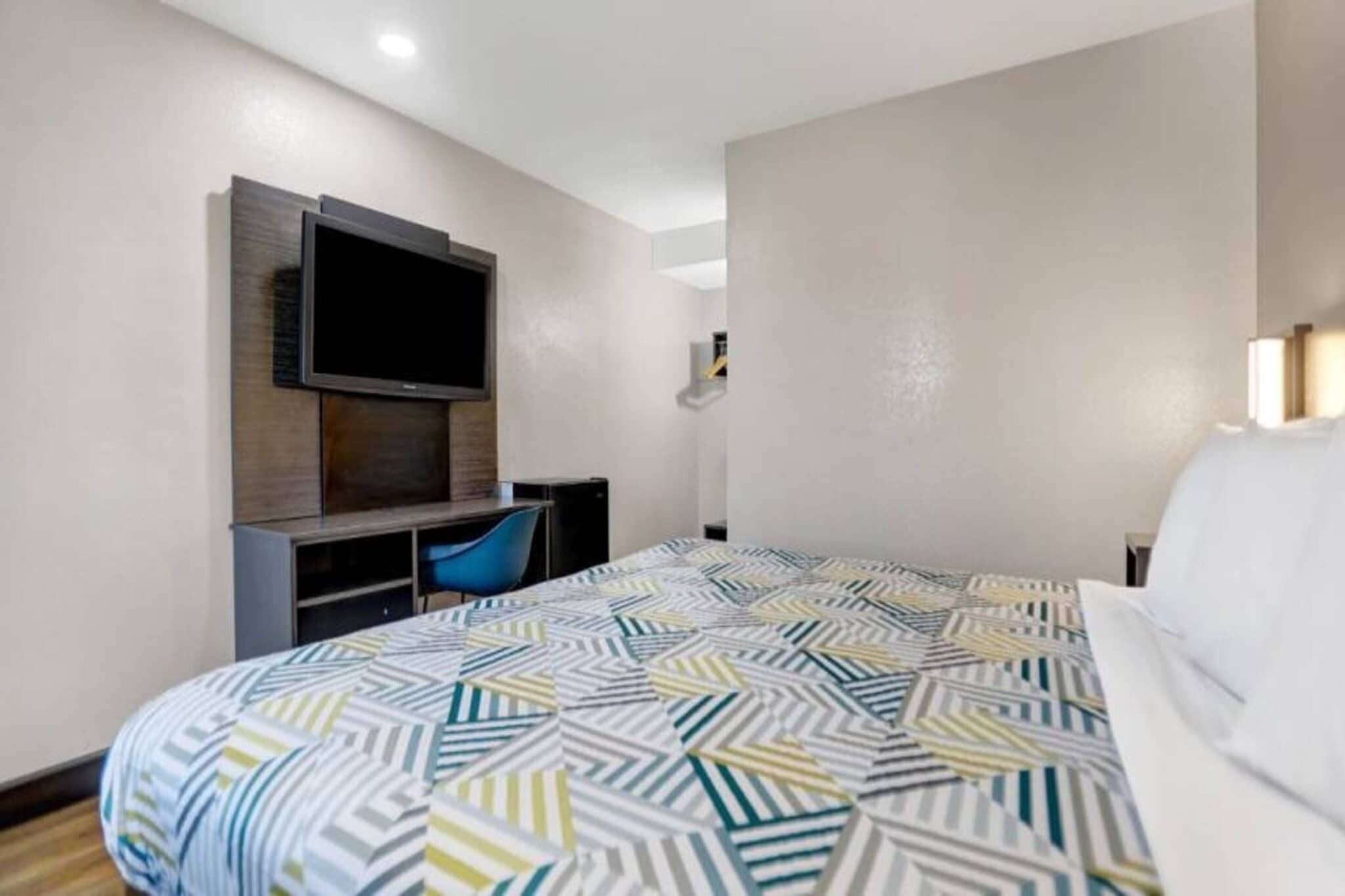 A hotel room with a bed, a desk with a chair, a flat-screen TV mounted on the wall, and a small table.