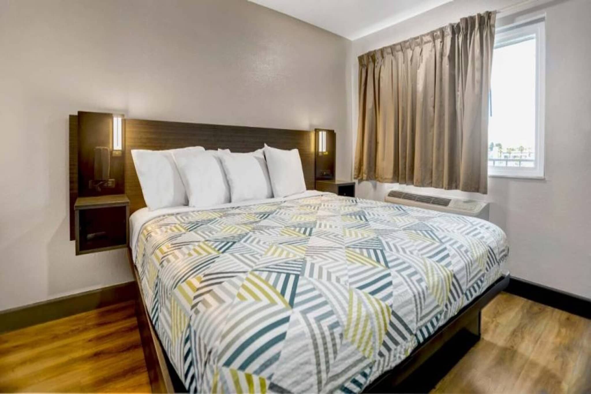 A hotel room with a bed, a nightstand, a wall-mounted lamp, a window with curtains, and a radiator.