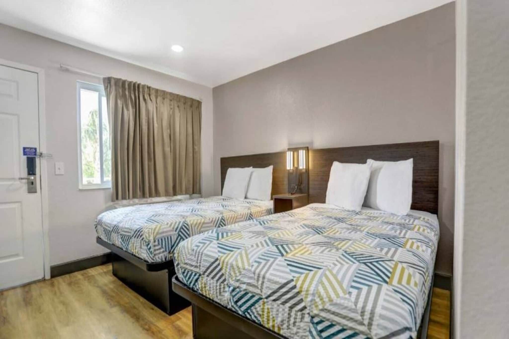 A hotel room with two beds, a window with curtains, a door, and a nightstand with a lamp.
