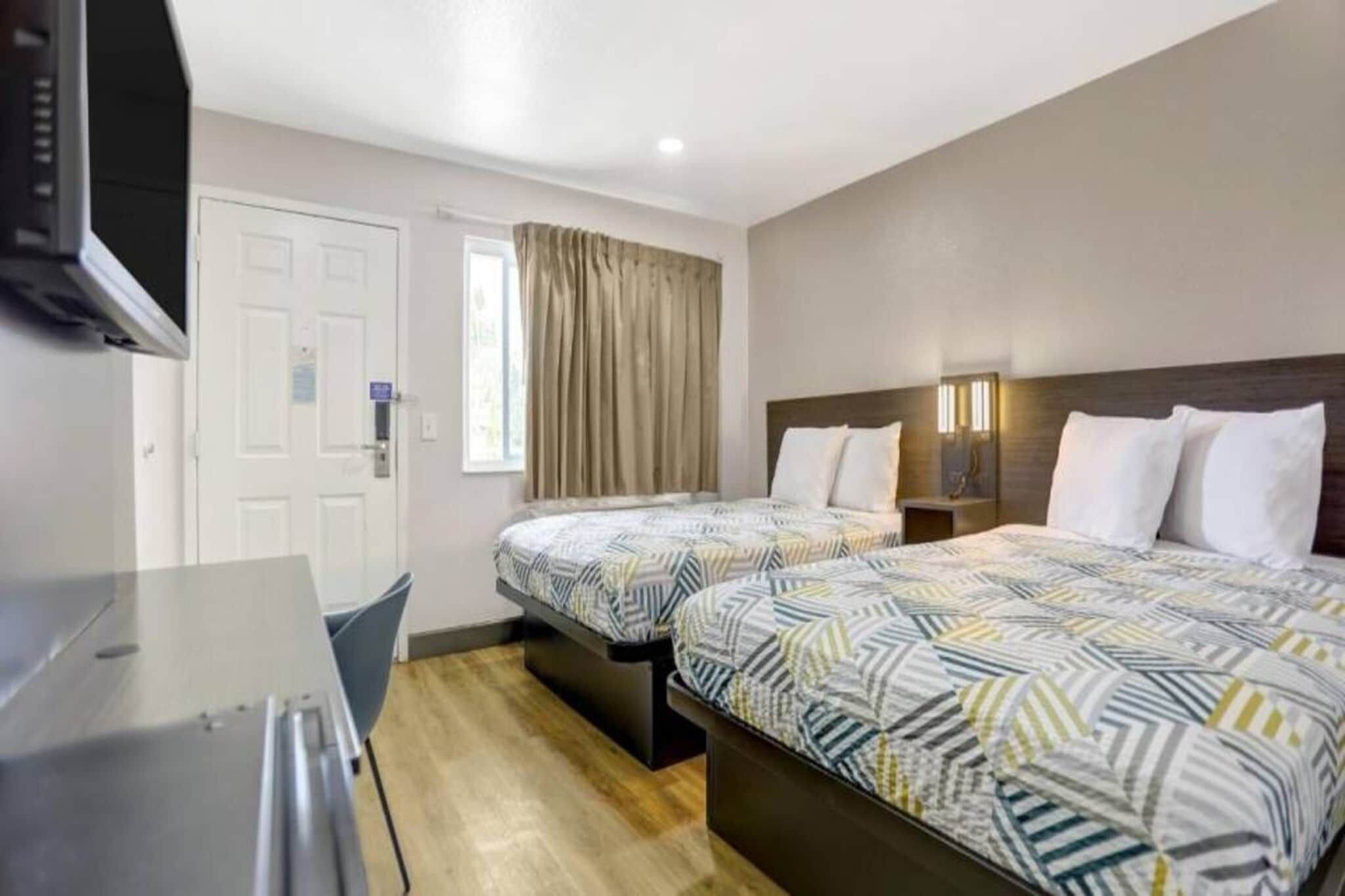 A hotel room with two beds, a desk, a television, and a window with curtains.
