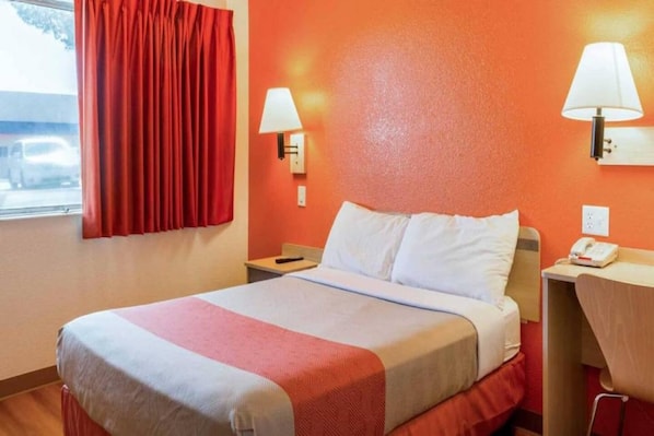 A hotel room with a bed, a nightstand, a desk, a chair, and a window with curtains.