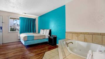 A hotel room with a bed, a small bedside table, a window with curtains, a bathroom with a bathtub, and a tiled floor.