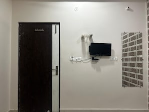 A room with a closed door, a mounted flat-screen TV, a wall-mounted light switch, and a decorative tiled wall. - Hotel Kashi Varanasi (varanasi)