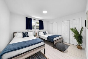 A bedroom with two beds, a window with curtains, a potted plant, and a door. - 5 Lakeside Cottages + Private Patios | Near Vanderbilt Beach | Loblolly Cottages (Naples)