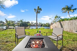 An outdoor seating area with a fire feature, two chairs, and a view of a lake and palm trees. - 5 Lakeside Cottages + Private Patios | Near Vanderbilt Beach | Loblolly Cottages (Naples)