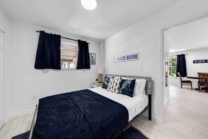 Een slaapkamer met een bed, nachtkastje, raam met gordijnen en een bordje met de tekst 'BEACH HOUSE'.
