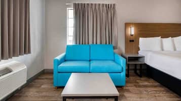 A hotel room with a blue sofa, a white bed, a small table, and a window with curtains.