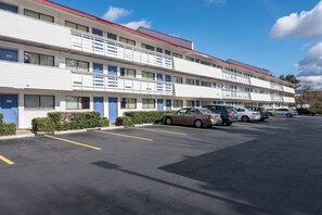 Un motel de varios pisos, cada uno con una puerta azul y número de habitación. Hay un estacionamiento con varios coches aparcados.