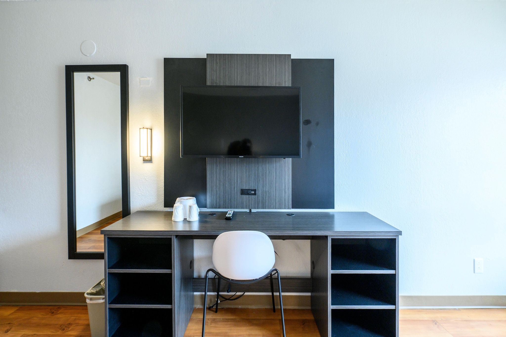 A modern hotel room with a dark wood desk, a white chair, a large mirror, and a flat-screen TV mounted on the wall.