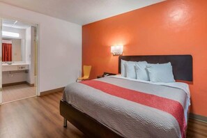 Une chambre d’hôtel avec un lit, une table de chevet, une chaise, une salle de bain avec un miroir et un mur de couleur orange.