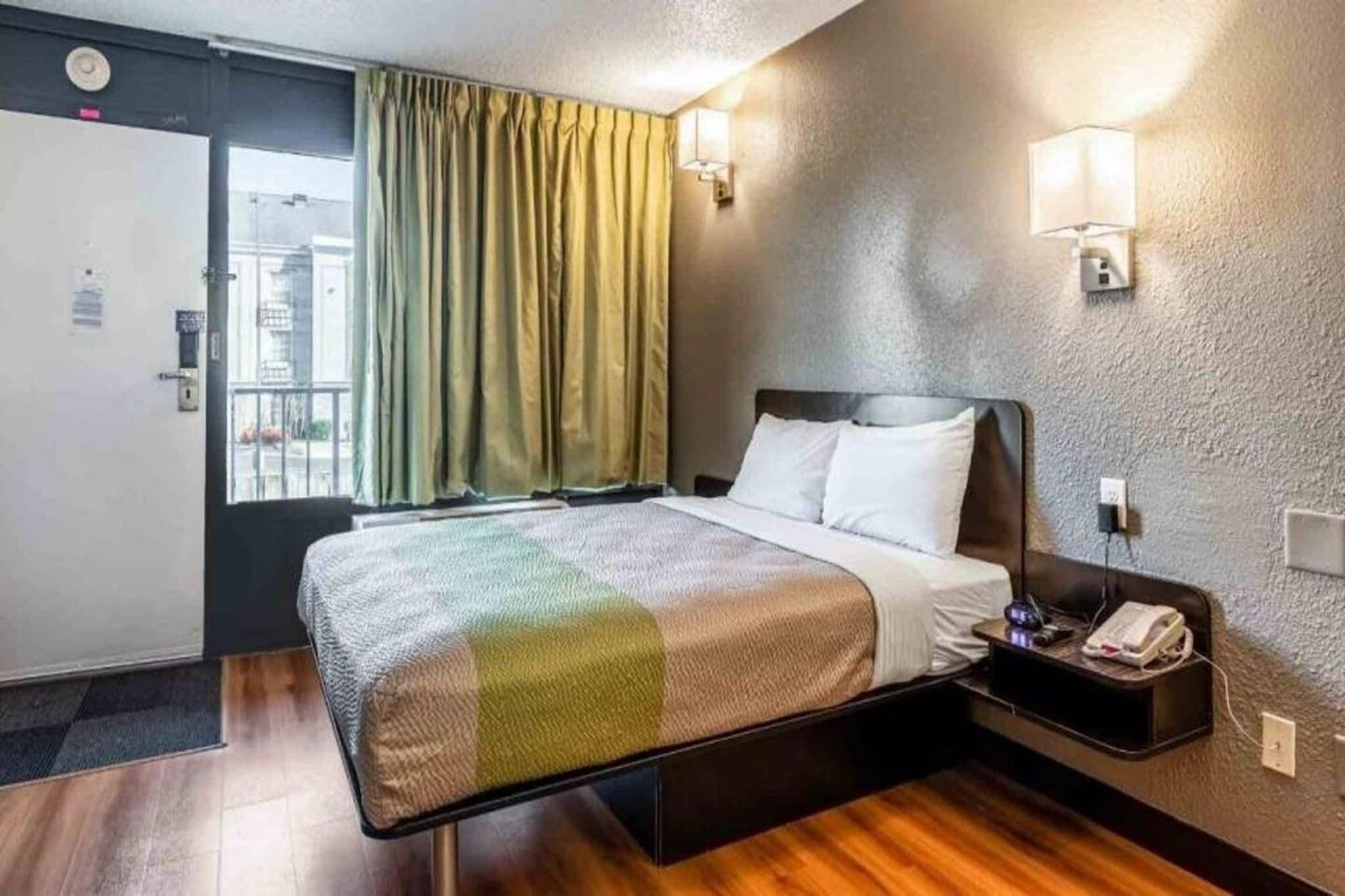 A hotel room with a bed, a nightstand, a lamp, and a window with curtains.