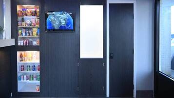 A room with a vending machine stocked with snacks and drinks, a television mounted on the wall, and a door.