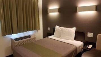 Quarto de hotel com cama, mesa de cabeceira, luminária, ar-condicionado na parede e janela com cortinas.
