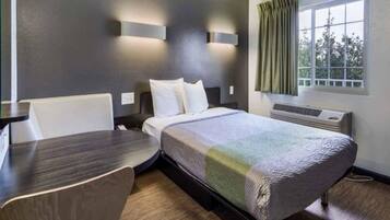 A hotel room with a bed, a desk, a chair, a window with curtains, and an air conditioning unit.