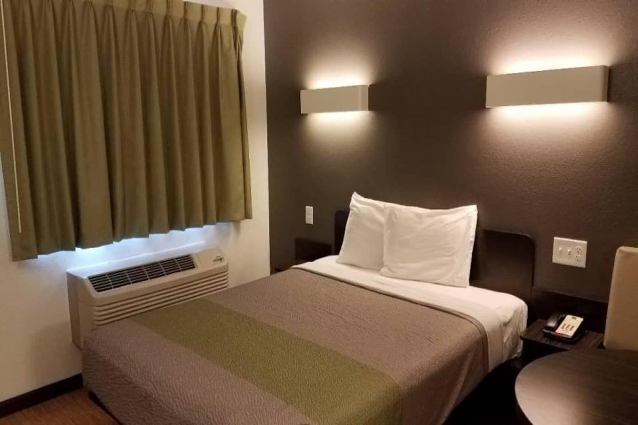 A hotel room with a bed, a bedside table, a lamp, a wall-mounted air conditioner, and a window with curtains.