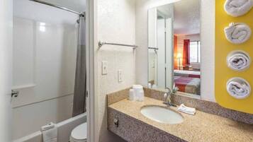 Baño con ducha, inodoro, lavamanos y espejo que refleja una cama y ventana.