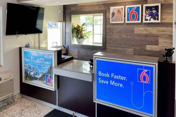 A reception desk with a digital screen displaying a promotional message, a television mounted on the wall, and framed pictures on the wall.