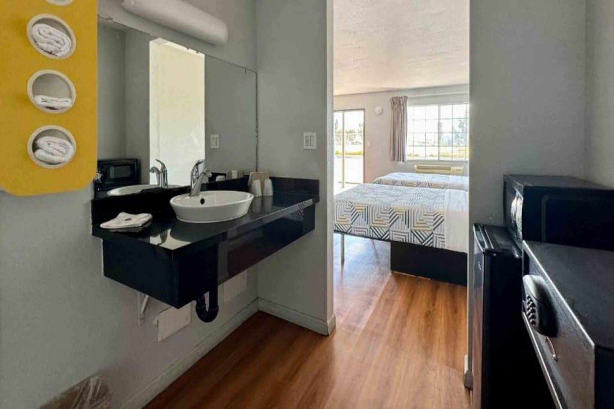 A hotel room with a bathroom featuring a sink, mirror, and towel dispenser. The room has a bed with a patterned bedspread and a television.