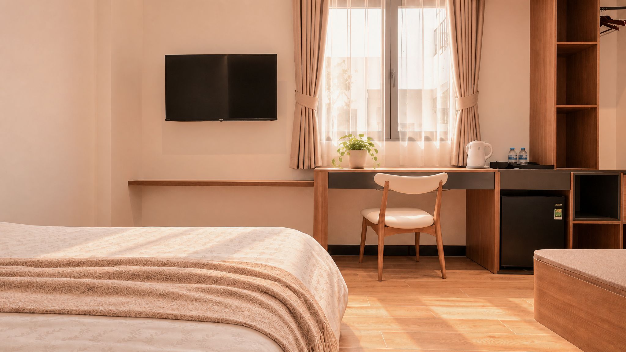 Habitación de hotel con cama, escritorio con silla, televisor, ventana con cortinas, planta pequeña y refrigerador.
