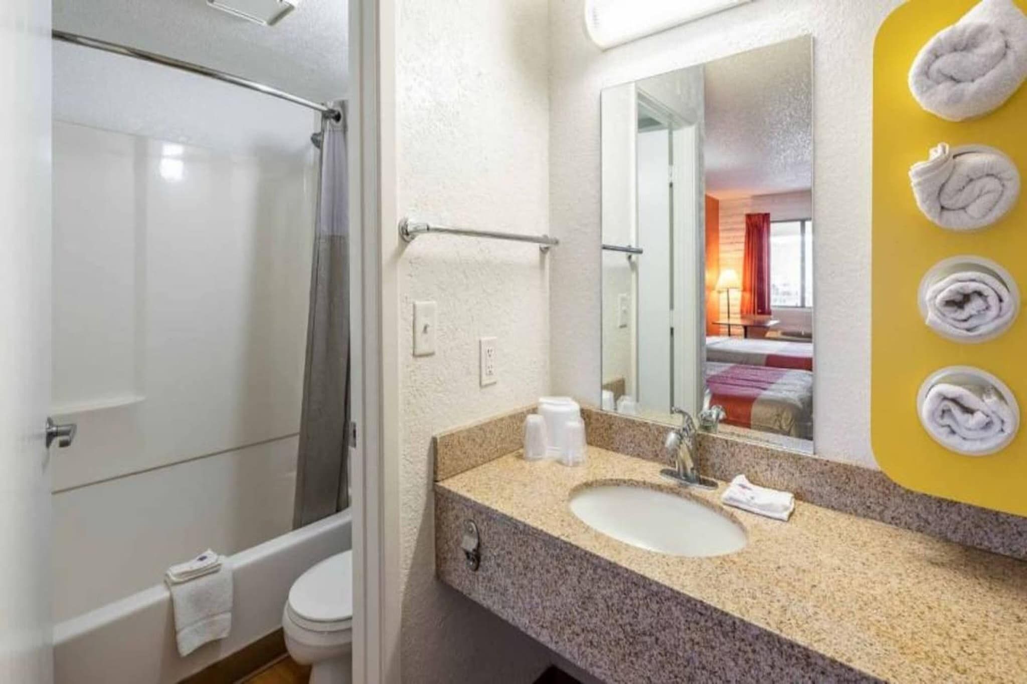 A bathroom with a shower, toilet, sink, and a mirror reflecting a bed and window.
