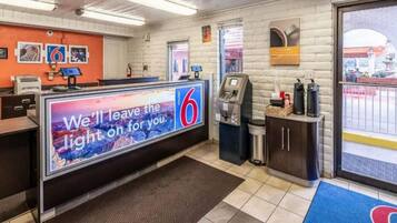 Een motel lobby met een receptiebalie, een digitaal bord, een koffieapparaat en uitzicht op de parkeerplaats.