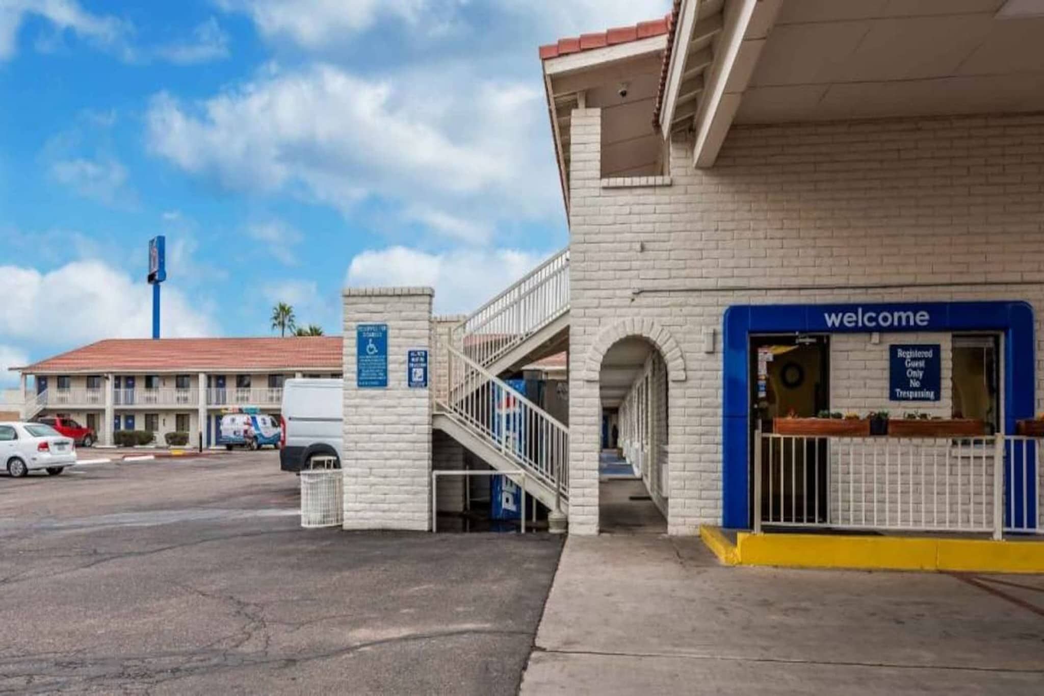 A hotel entrance with a 'Welcome' sign, a ramp for accessibility, and a parking area with cars.