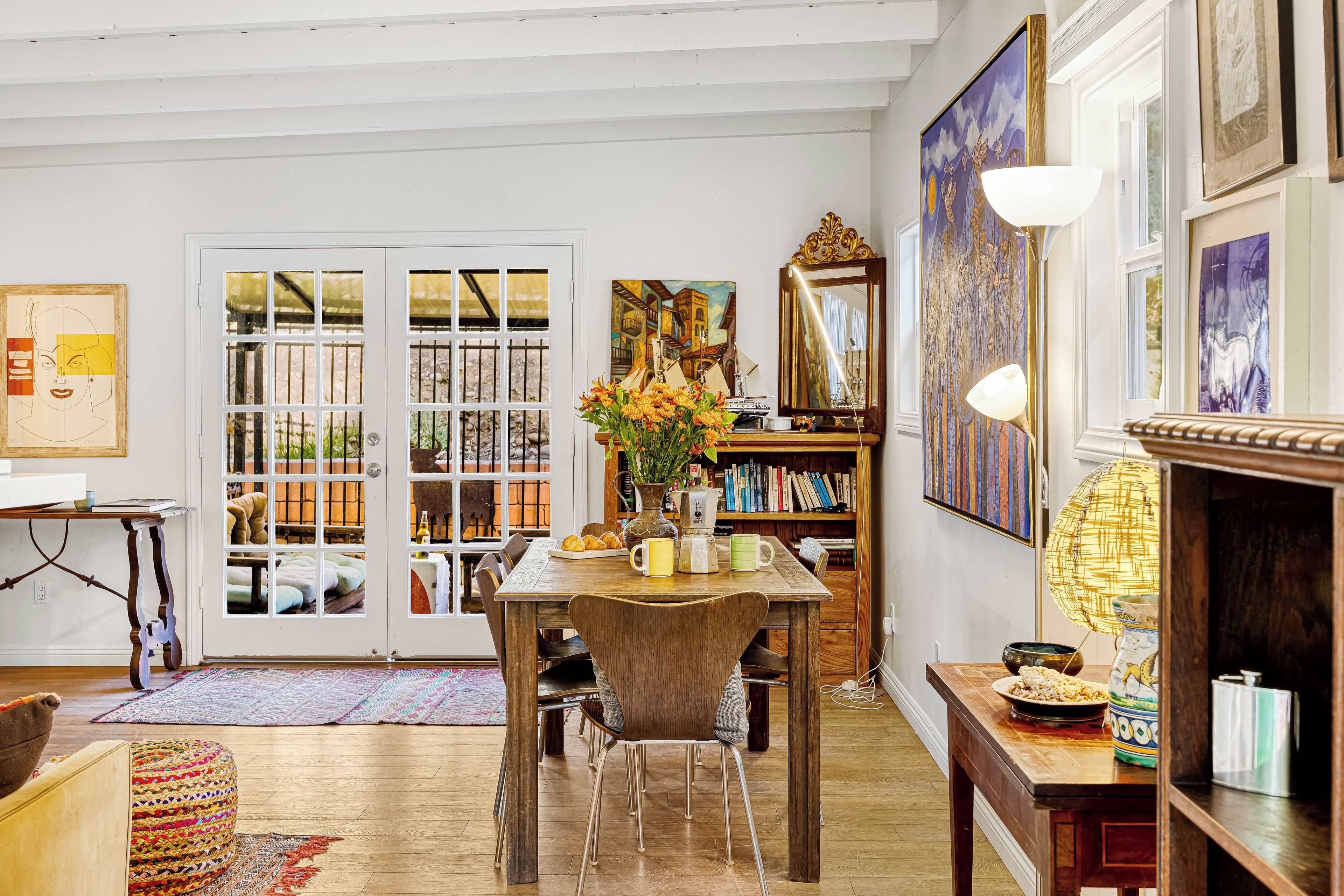 A room with a wooden dining table, chairs, a bookshelf, and a large window with a view of the outside.