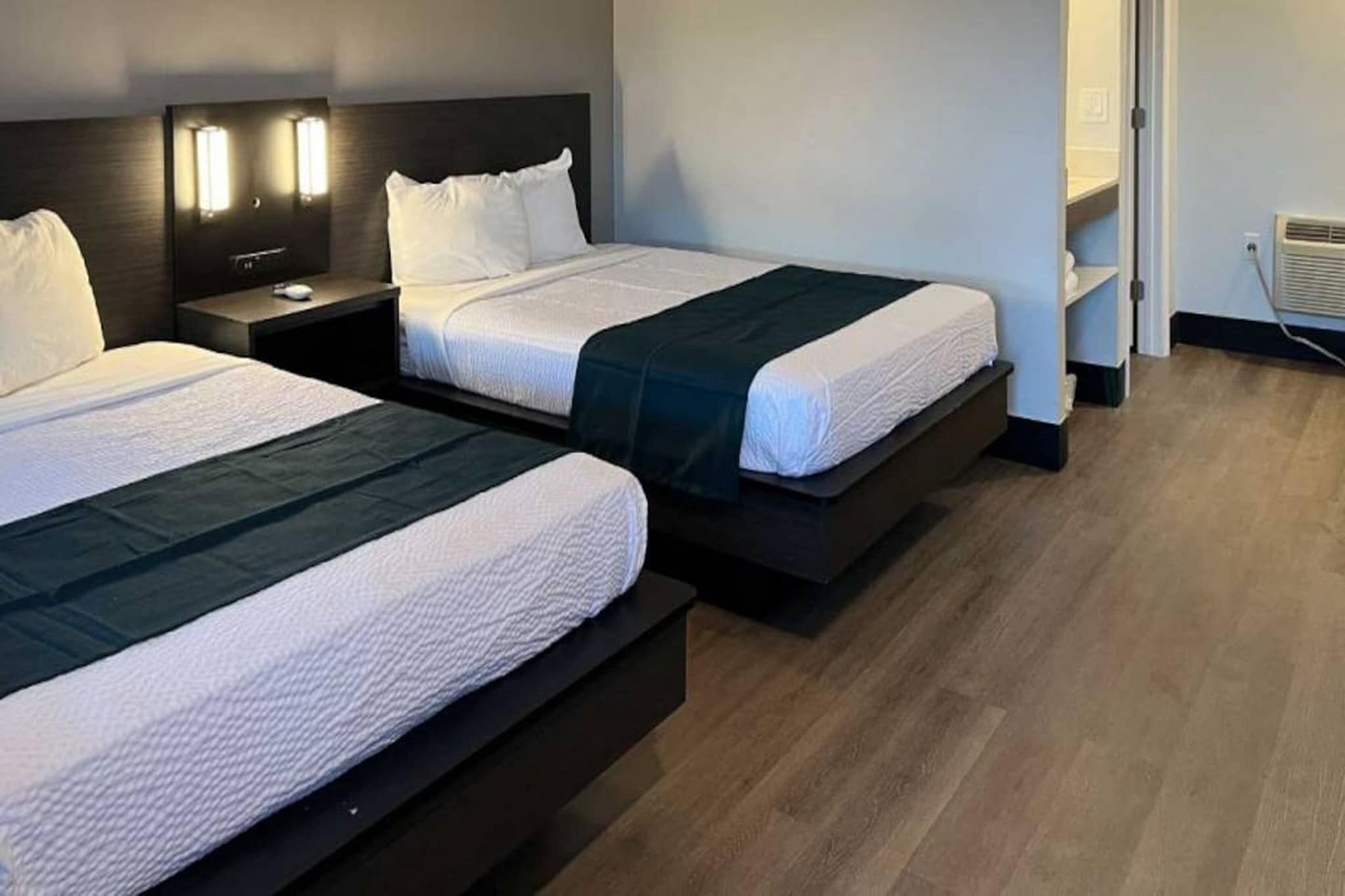 Ein Hotelzimmer mit zwei Betten, beide mit dunkelbraunen Kopfteilen und weißer Bettwäsche. An jedem Bett befindet sich ein Nachttisch mit einer Lampe und ein kleiner Tisch. Der Raum hat Holzböden und eine Tür, die in einen weiteren Bereich führt.