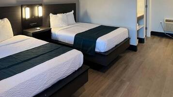 Ein Hotelzimmer mit zwei Betten, beide mit dunkelbraunen Kopfteilen und weißer Bettwäsche. An jedem Bett befindet sich ein Nachttisch mit einer Lampe und ein kleiner Tisch. Der Raum hat Holzböden und eine Tür, die in einen weiteren Bereich führt.