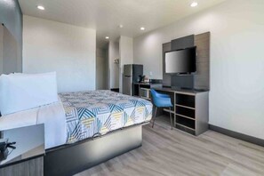 A modern hotel room with a bed, a desk, a chair, a television, and a kitchenette.