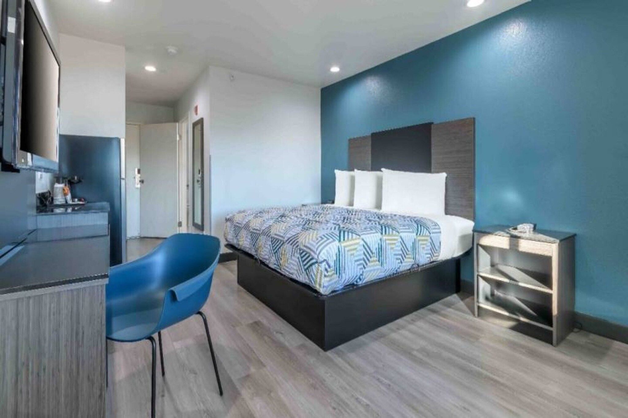 A modern hotel room with a bed, a desk, a chair, and a small bedside table.