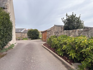 Un carrer pavimentat estret, amb murs de pedra i vegetació als costats, que condueix a un edifici de fusta amb teulada inclinada.