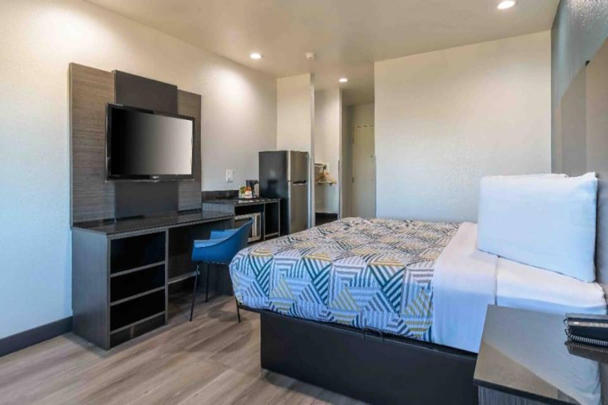 A modern hotel room with a bed, a desk, a chair, a television, and a wardrobe.