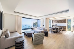 Ein modernes Wohnzimmer mit einer Couch, einem Couchtisch und einem gemusterten Ottomanen. Es gibt eine Essecke mit einem runden Tisch und Stühlen. Das Zimmer hat große Fenster mit Meerblick.