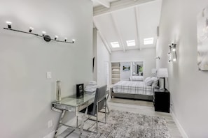 Une chambre à coucher moderne avec un bureau en verre, un lit recouvert d’une couverture à carreaux et une lucarne au-dessus.